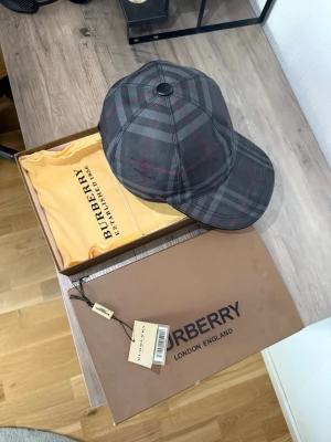 Burberry  keps - Snygg keps från Burberry i klassiskt rutmönster i grått, svart och röda detaljer. Justerbar rem bak och böjd skärm. Tillverkad i bomull och polyester. Perfekt accessoar för dig som vill ha en stilren och exklusiv look. Använd ca 2-3 gånger max pga att den är lite stor för mig. Kommer även med box,  etikett samt en påse till kepsen av burberry.