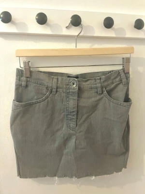 Grå kort jeanskjol med rå kant - Säljer en grå kort jeanskjol med rå, fransig kant nertill.  På bakfickorna finns broderade detaljer med små nitar som ger extra stil. Perfekt till sommaren. Kjolen är självsydd men som xs. 