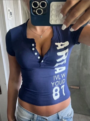 Mörkblå croppad topp med tryck - Snygg mörkblå croppad topp med vit text och siffror framtill. Toppen har korta ärmar, djup v-ringning med knappar och figurnära passform. Perfekt till jeans för en trendig look.