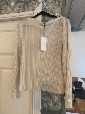 Beige plisserad långärmad topp - Trendig beige topp med plisserad struktur och lång ärm. Toppen har en rak passform och är tillverkad i ett lätt, syntetiskt material som ger en snygg textur. Perfekt för dig som gillar stilrena och moderna plagg.