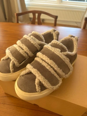 Grå sneakers med fårull och kardborre - Unika sneakers från Inuikii i grå mocka med fluffiga detaljer av fårull och breda kardborreband. Skorna har en chunky vit sula och en mysig insida, perfekta för dig som vill sticka ut med din stil.