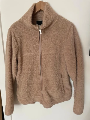 Beige teddyjacka med dragkedja - Mysig beige teddyjacka med hög krage och hel dragkedja framtill. Jackan har två sidofickor och är gjord i ett fluffigt, mjukt material🧡