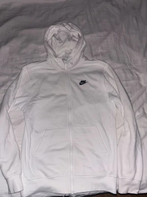 Vit hoodie från Nike med dragkedja - Snygg vit hoodie från Nike med dragkedja framtill och klassisk huva. Har två stora fickor på magen och en liten svart Nike-logga broderad på bröstet. Perfekt för en avslappnad och sportig stil. Använd fåtal gånger. Sitter kompakt och samtidigt skönt. 
