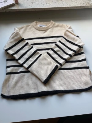 Beige stickad tröja med svarta ränder - Snygg beige stickad tröja med svarta horisontella ränder och rund hals. Tröjan har långa ärmar och ribbade muddar samt en lös, avslappnad passform. Perfekt för lager på lager och enkel att styla till jeans eller kjol.