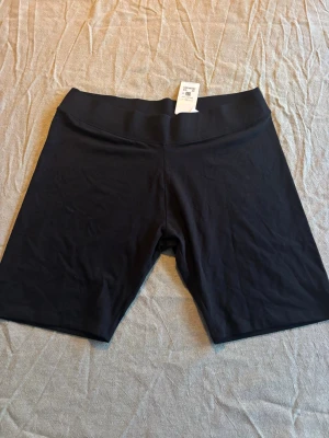 Ny med prislapp, essence bike shorts, svarta - Säljer ett par svarta cykelshorts med bred resår i midjan. De är midwaist och har en enkel, stilren design utan synliga detaljer. Perfekta för träning eller som basplagg till vardags. Materialet känns mjukt och stretchigt. Helt nya med prislapp kvar.