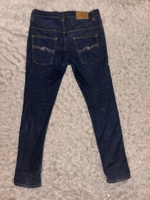 Nudie jeans - Säljer ett par mörkblå skinny jeans från Nudie med klassiska kontrastsömmar och broderade bakfickor. Jeansen har fem fickor och en smal passform som sitter snyggt. Perfekta till vardagslooken! Modellen är thinn fin