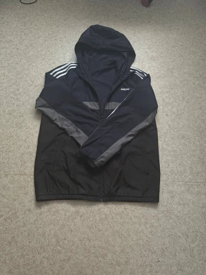 Svart vindjacka från Adidas - Snygg svart vindjacka från Adidas med huva och dragkedja. Jackan har vita ränder på axlarna och grå detaljer längs ärmarna. Tillverkad i lätt och tåligt material, perfekt för blåsiga dagar. Adidas-logga på bröstet ger en sportig vibe. TVÅ JACKOR I EN!