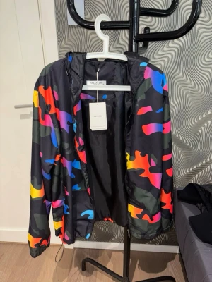 Färgglad camo vindjacka med huva - Säljer en riktigt snygg vindjacka med färgglatt camomönster i neonfärger på svart botten. Passar de som har storlek S/M, liten i storleken. Jackan har huva och dragkedja framtill. Perfekt för dig som vill sticka ut och gillar streetwear vibes. Materialet är tunt och smidigt, passar bra till lager på lager. Helt ny aldrig använd 