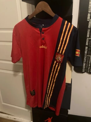 Spansk Adidas fotbollströja röd/blå - Säljer en röd och marinblå fotbollströja från Adidas med Spaniens landslagsemblem och gula ränder. Tröjan har korta ärmar, knappslå vid halsen och broderad Adidas-logga. Perfekt för dig som älskar fotboll och vill visa ditt stöd för Spanien.