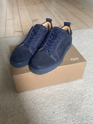 Mörkblåa loubs  - Snygga mörkblå sneakers i mocka med klassisk låg profil och matchande blå snörning. Skorna har en ikonisk röd sula med präglad text och beige innerfoder. Perfekta för dig som gillar stilrena detaljer och vill sticka ut.