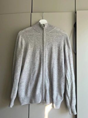 Ljusgrå ziptröja från Soft Goat - Ljusgrå ziptröja från Soft Goat i mjuk kashmir. Tröjan har hög krage, hel dragkedja framtill och ribbade muddar vid ärmslut och nederkant. Perfekt för lager på lager och en clean look.