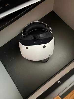 VR-headset med kontroller - Säljer ett stilrent VR-headset i vit och svart design med matchande kontroller. Headsetet har en rundad form och justerbart band för bekväm passform. Kontrollerna har en modern look och passar perfekt för gaming och uppslukande VR-upplevelser.