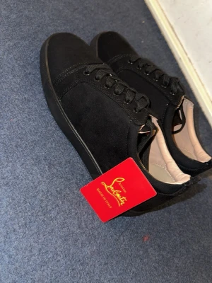 Svarta Louboutin sneakers med röd sula. Kommer utan box - Snygga svarta sneakers från Louboutin med klassisk röd sula och svarta skosnören. Skorna har en låg profil och är tillverkade i mocka med detaljer i läder. Perfekta för dig som vill sticka ut med en exklusiv touch.