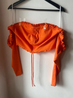 Orange offshoulder croppad topp - Trendig orange offshoulder topp med långa ärmar och croppad passform. Toppen har volangkant upptill och snörning framtill för en snygg detalj. Perfekt för dig som vill sticka ut med färg och stil.