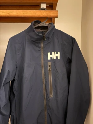 Marinblå vindjacka från Helly Hansen - Snygg marinblå vindjacka från Helly Hansen med hög krage och dragkedja framtill. Jackan har en bröstficka med dragkedja och HH-logga på bröstet. Tillverkad i lätt och slitstarkt material, perfekt för blåsiga dagar.