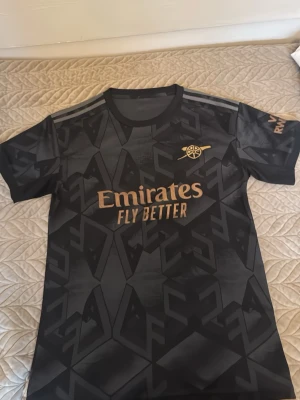  Arsenal fotbollströja - Snygg svart Arsenal t-shirt med geometriskt mönster och guldfärgade detaljer. Tryck med 'Emirates Fly Better' på bröstet och klubbens logga. T-shirten har rund hals och korta ärmar, perfekt för dig som gillar fotboll och vill ha en sportig look.