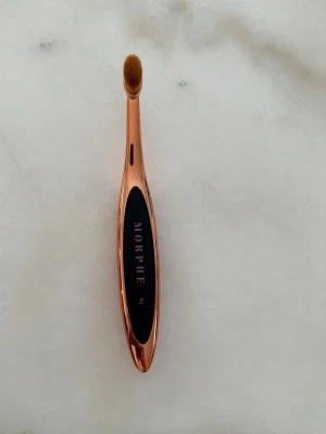 Oanvänd sminkborste från Morphe - M6 Nose Contour Brush från det Morphe x Brittany Bear 360 Nose Contour Collection-samarbetet. Design: Ergonomiskt utformat handtag i roséguld med svart mittsektion och Morphe M6-märkning.