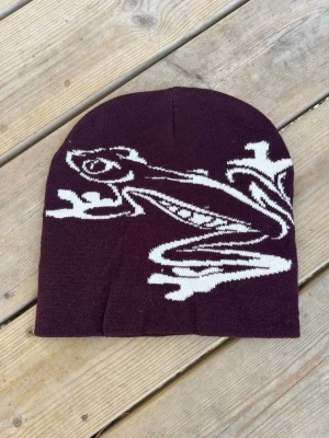 Lila mössa med grodmotiv - Säljer en lila mössa med ett stort vitt grodmotiv på framsidan. Mössan är stickad och har en klassisk, rundad form som sitter skönt på huvudet. Perfekt för dig som vill sticka ut med en unik accessoar.