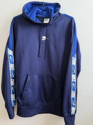 Blå Nike hoodie med logga på ärmar - Mörkblå hoodie från Nike med huva och känguruficka. Ärmarna har blå och vita band med Nike-loggor längs sidorna. Klassisk Nike-logga på bröstet och dragsko i huvan. Perfekt för en sportig och avslappnad stil. Passar även S 