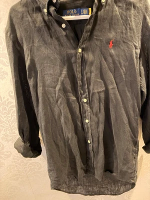 Svart linneskjorta från Polo Ralph Lauren - Snygg svart skjorta i linne från Polo Ralph Lauren med klassisk custom fit. Skjortan har långa ärmar, knappar framtill och den ikoniska röda logotypen broderad på bröstet. Perfekt för dig som gillar stilrena och tidlösa plagg.