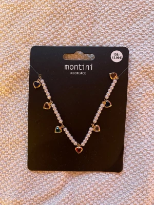 Pärlhalsband med färgglada hjärtan - Snyggt halsband från Montini med vita pärlor och små hjärtformade hängen i olika färger. Hjärtana har guldfärgade kanter och ger halsbandet en lekfull och trendig look. Perfekt accessoar för att lyfta vilken outfit som helst.