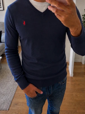 Mörkblå tröja Ralph Lauren - Mörkblå tröja från Polo Ralph Lauren med röd broderad logga på bröstet. Tröjan har långa ärmar, ribbade muddar och är tillverkad i ett mjukt material som passar perfekt till jeans.