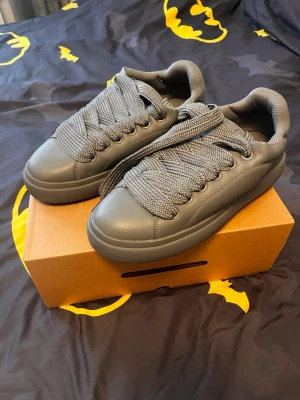 Bershka chunky sneakers med breda snören - Säljer ett par grå sneakers med breda snören och chunky sula. Skorna har en clean och modern look med rund tå och är tillverkade i ett syntetmaterial som ger en slät finish. Perfekta för dig som gillar statement-detaljer på dina skor.