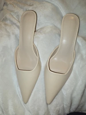 Kitten heels mules - strl 38 - Helt ny skick. Snygga pumps i beige med spetsig tå och öppen häl. Skorna har en smal klack och en stilren, minimalistisk design. Tillverkade i ett slätt material som ger en elegant look. Perfekta för dig som gillar trendiga och klassiska skor.