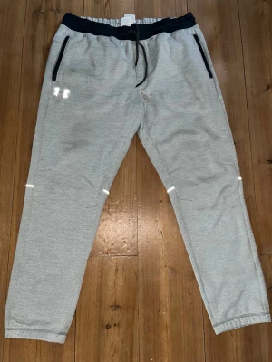 Grå mjukisbyxor från Under Armour - Snygga grå mjukisbyxor från Under Armour med svarta detaljer och dragsko i midjan. Byxorna har två fickor med dragkedja och elastisk midja för extra komfort. Perfekta för chill eller träning. Endast använd ett fåtal gånger på gym! Nypris:549kr.  Mitt pris:320