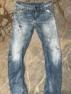  G-Star Jeans - Säljer ett par ljusblå jeans från G-Star med slitningar och hål på både fram- och baksida. Jeansen har fem fickor, knappgylf och en relaxed fit. Materialet är klassisk denim i bomull och de har en cool, tvättad look.