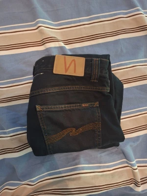 Mörkblå jeans från Nudie Jeans - Säljer ett par stilrena svarta Nudie Jeans i storlek W30/L32. Jeansen är i bra begagnat skick med normalt slitage och passar perfekt till både vardag och fest. Bekväm passform och en tidlös modell. (Lappen i byxorna saknas)