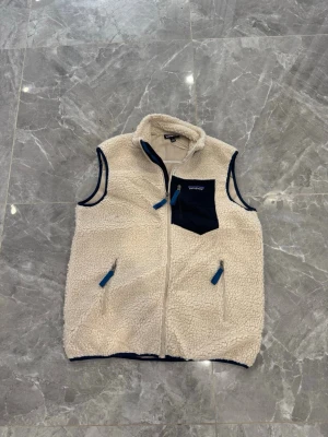 Beige fleeceväst från Patagonia - Mysig beige fleeceväst från Patagonia med marinblå detaljer och dragkedjor. Västen har en hög krage, två sidofickor och en bröstficka i kontrasterande tyg med logga. Perfekt för lager-på-lager och outdoor-vibbar.
