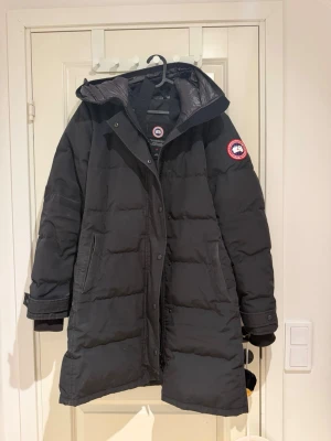 Svart lång vinterjacka Canada Goose - Säljer en svart lång dunjacka från Canada Goose med huva och klassisk logga på ärmen. Jackan har två stora fickor framtill, dragkedja och knappar samt justerbara muddar. Perfekt för kalla vinterdagar och riktigt varm. Jackan behöver bara en tvätt.