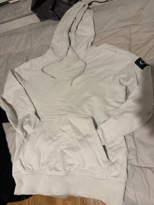 Vit hoodie från Calvin Klein Jeans - Snygg vit hoodie från Calvin Klein Jeans med stor magficka och dragsko i huvan. Klassisk passform och ribbade muddar. Liten patch på ena ärmen för extra detalj. Perfekt för en clean och stilren look.