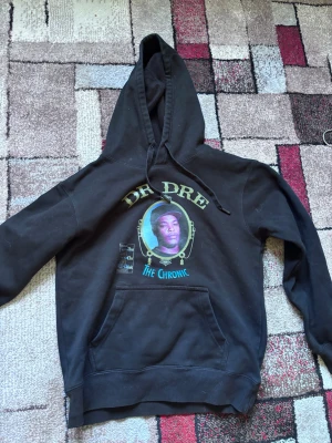 Svart Dr. Dre The Chronic hoodie - Säljer en svart hoodie med stor huva och magficka. På framsidan finns ett färgglatt tryck med Dr. Dre och texten 'The Chronic'. Hoodien har ribbade muddar och är tillverkad i mjukt bomullsmaterial. Perfekt för dig som gillar hiphop och streetwear.