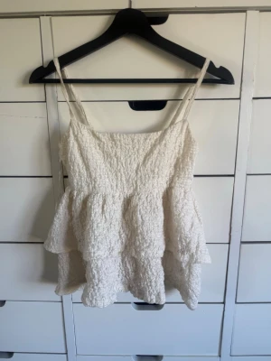Vit volang linnetopp med struktur - Supersöt vit linnetopp med smala axelband och volangdetaljer. Toppen har en strukturerad, lite skrynklig yta som ger en unik look. Perfekt för varma dagar och enkel att styla med jeans eller kjol.