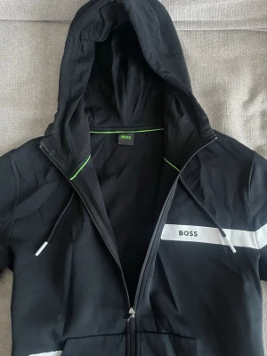Hugo zip hoodie svart med vita detaljer  - Säljer denna snygga zip hoodie då den inte kommer till användning längre. Storlek L och sitter som en L. Använt fåtal gånger cirka 1-3 gånger men den ser helt ny ut. Kan gå ner i pris i en snabb och smidig affär! Köparen står för frakten😁