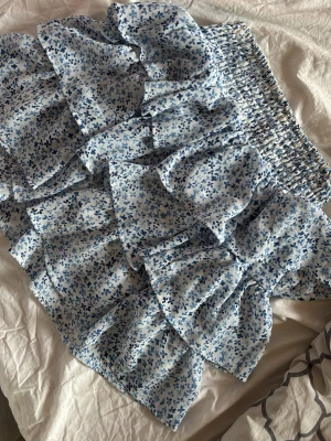 Blommig volangkjol i blått och vitt - Supersöt kort volangkjol med blått och vitt blommönster. Kjolen har bred smock i midjan för skön passform och flera lager volanger som ger en luftig och somrig känsla. Knappt använd