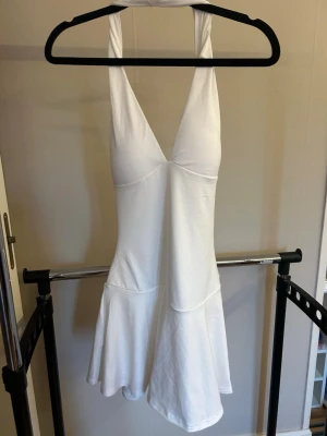 Vit kort halterneckklänning - Säljer en vit kort klänning med halterneck och djup v-ringning framtill. Klänningen har en öppen rygg och lätt utsvängd kjol. Perfekt för dig som vill ha en enkel men snygg look till sommaren. Aldrig använd