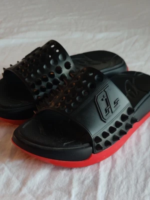 Svarta sandaler med nitar och röd sula - Coola svarta sandaler med bred rem dekorerad med svarta nitar och ett präglat mönster. Sulan är svart med en röd undersida för en extra edgy look. Tillverkade i syntetmaterial och har en sportig vibe med unika detaljer.