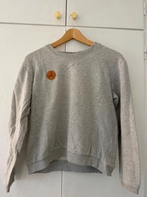 Grå sweatshirt med rund hals - Enkel och stilren grå sweatshirt med rund halsringning och ribbade muddar. Tröjan har ett litet brunt märke med djurmotiv på bröstet. Perfekt basplagg i mjukt material och avslappnad passform.