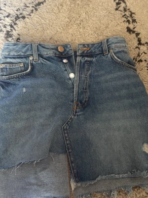 Blå jeanskjol med fransig kant - Säljer en blå jeanskjol med rå, fransig nederkant och slitna detaljer. Kjolen har klassisk femficksdesign, knappgylf och hög midja. Perfekt för dig som gillar en avslappnad och trendig stil.