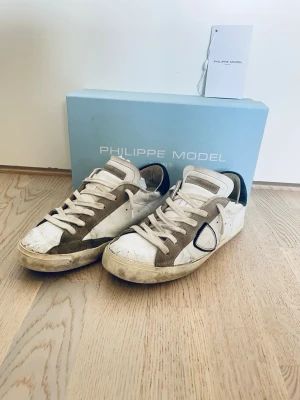 Philippe Model vita sneakers - Snygga vita sneakers från Philippe Model med beige mockadetaljer och svarta partier vid hälen. Klassisk låg modell med vita snören och ikonisk logga på sidan. Skorna har en cool vintage-look och passar perfekt till streetwear eller casual outfits. De är ganska slitna men själv så tycker jag att det gör de snyggare. Sulan är dock i sisådär skick men det är väldigt vanligt på Philippe model sneakers och svårt att undvika. Priset är diskuterbart!