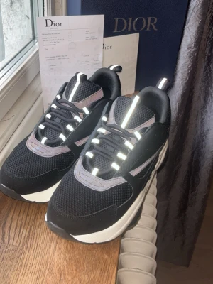 Dior B22 Storlek 43 - Snygga svarta sneakers från Dior med vita och grå detaljer samt reflexdetaljer på snörningen och tungan. Ovandelen är i mesh och läder, vilket ger både stil och komfort. Tjock vit sula för en modern look. Perfekta för dig som vill sticka ut.