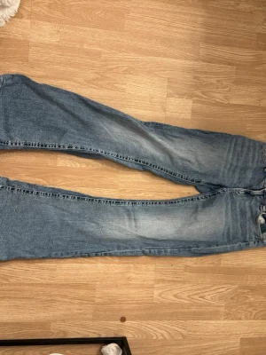Blå bootcut jeans med fem fickor - Säljer ett par klassiska blå jeans med rak passform och fem fickor. Jeansen har normalhög midja och subtila slitningar framtill för en avslappnad look. Tillverkade i denim med snygg tvättad effekt. Perfekta till vardagsstilen!