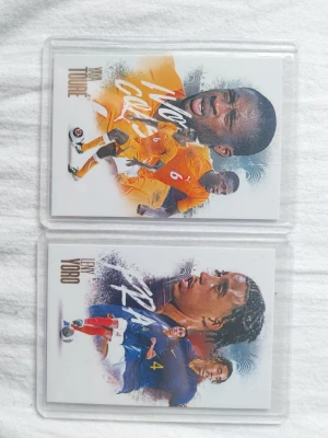 Fotbollssamlarbilder Yaya Touré & Leny Yoro - Två samlarbilder med fotbollsspelarna Yaya Touré och Leny Yoro. Korten har färgstarka illustrationer, en med orange matchställ och en med blått. Båda korten ligger i plastfickor och har spelarnas namn tryckt på framsidan.