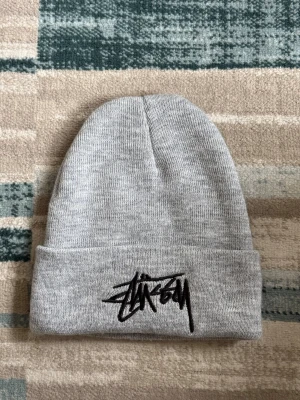 Grå Stüssy mössa med logga - Säljer en stilren grå mössa från Stüssy med svart broderad logga framtill. Mössan är ribbstickad och har en uppvikt kant för extra komfort. Perfekt accessoar för att hålla värmen och addera streetwear-vibe till din outfit.