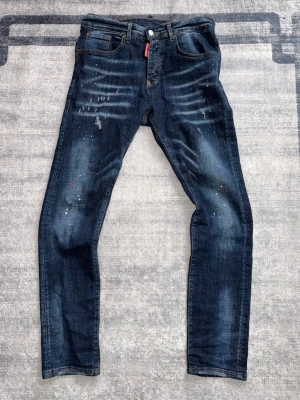 Mörkblå skinny jeans med slitningar - Mörkblå skinny jeans från Dsquared2 med slitningar, färgstänk och lätt tvättad look. Klassisk femficksmodell med läderpatch bak i midjan och röda detaljer vid gylfen. Perfekta för en trendig och avslappnad stil.