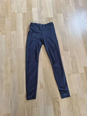 Svarta höga tränings leggings med ficka - Snygga svarta leggings med hög midja och praktiska sidoficka. Tillverkade i ett stretchigt och mjukt material som sitter skönt på kroppen. Perfekta för en sportig look eller till vardagsoutfiten.