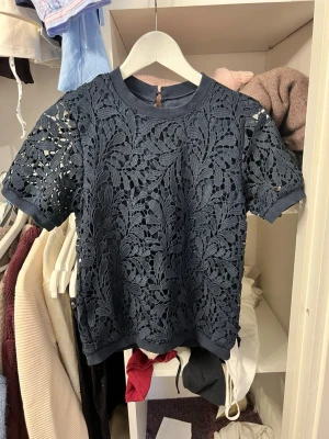 Mörkblå spetsblus med kort ärm - Snygg mörkblå blus i spets med korta ärmar och rund hals. Blusen har ett detaljerat blommönster och knapp i nacken. 